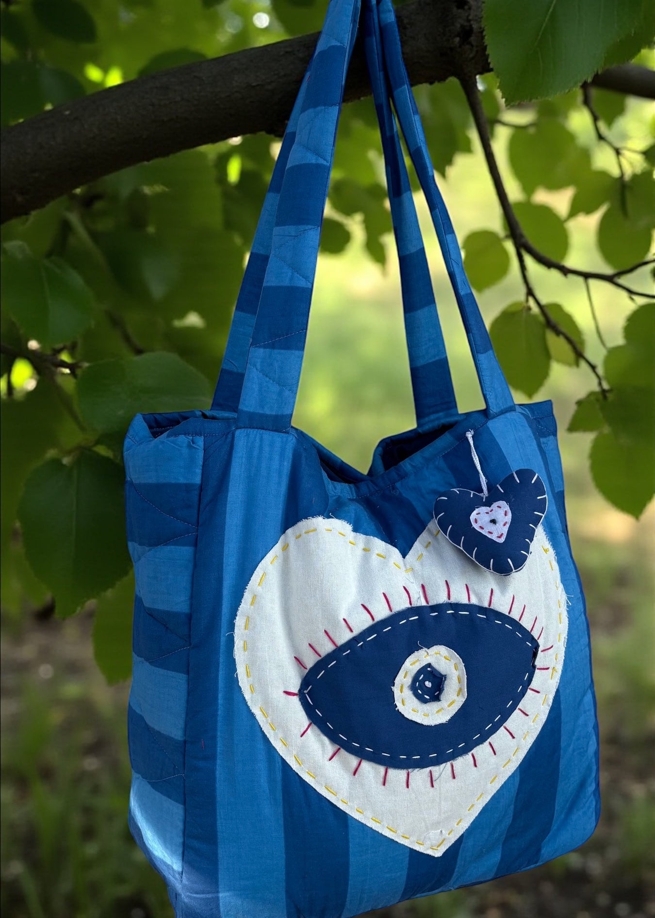 Blue striped quilted tote bag with heart-shaped evil-eye appliqué and matching padded charm hanging outdoors.