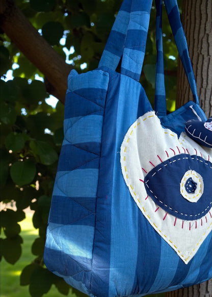 Blue striped quilted tote bag with heart-shaped evil-eye appliqué and matching padded charm hanging outdoors.