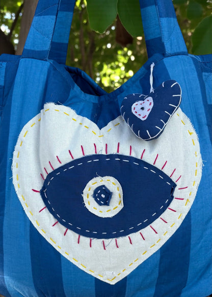 Blue striped quilted tote bag with heart-shaped evil-eye appliqué and matching padded charm hanging outdoors.
