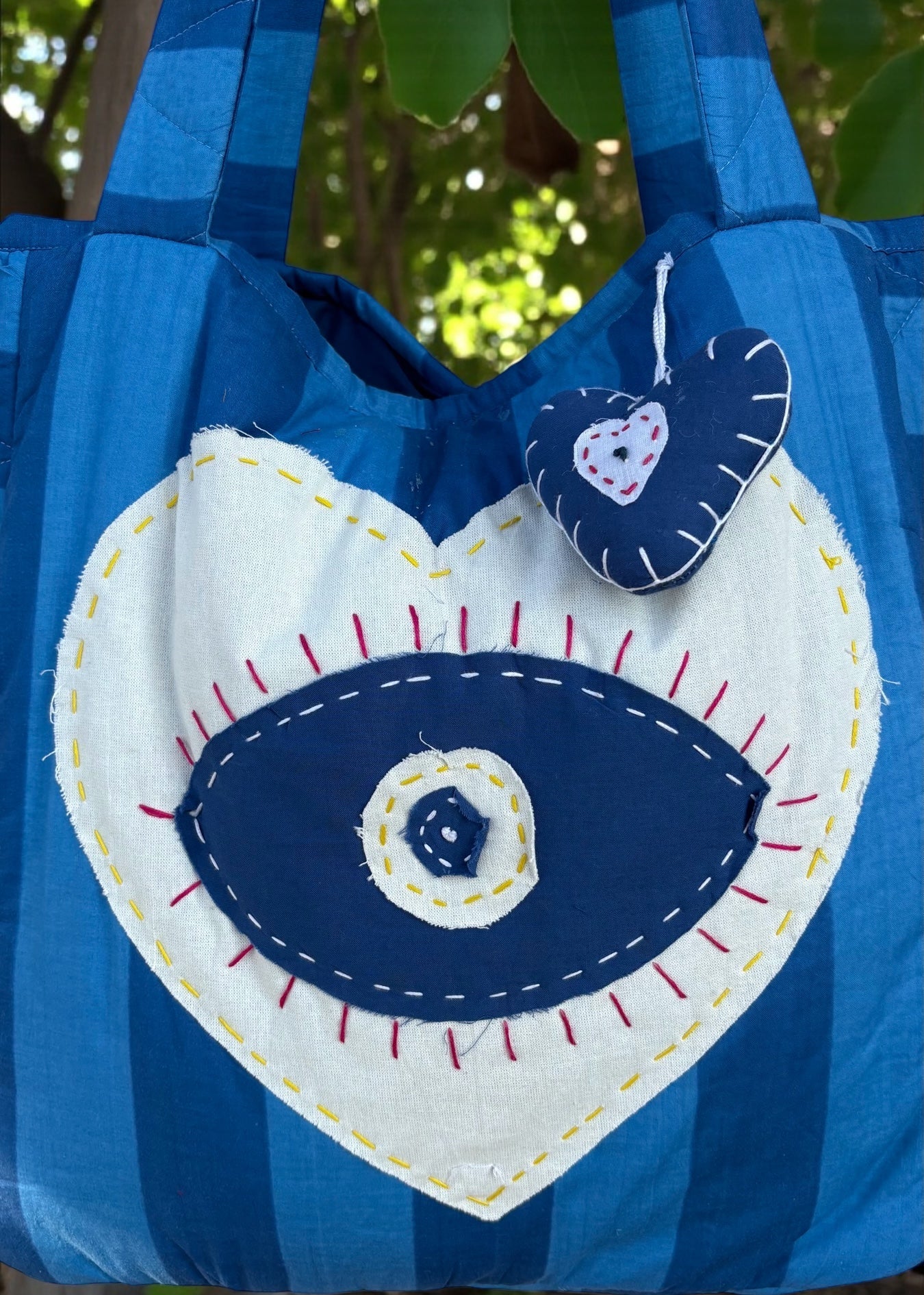Blue striped quilted tote bag with heart-shaped evil-eye appliqué and matching padded charm hanging outdoors.