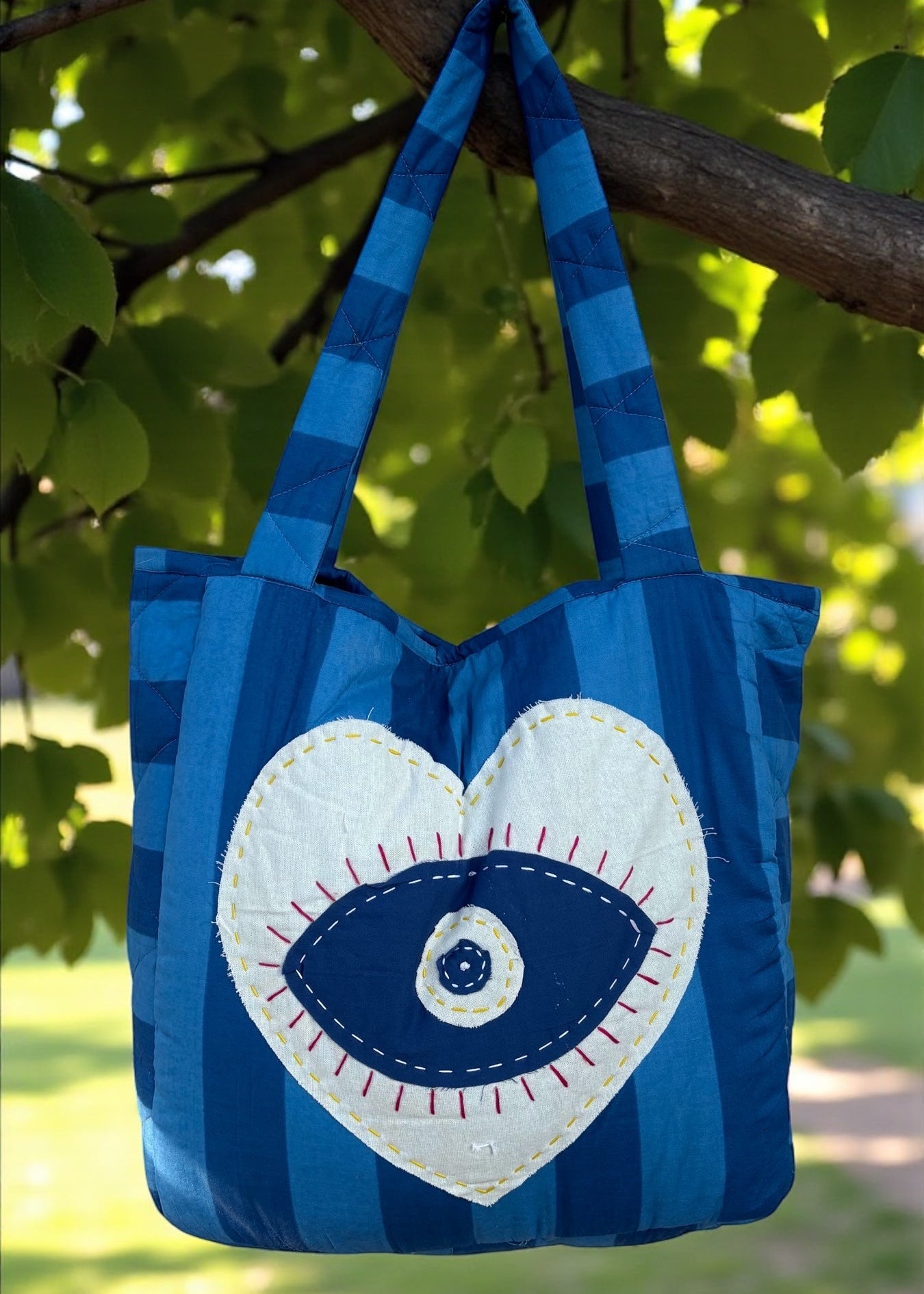 Blue striped quilted tote bag with heart-shaped evil-eye appliqué and matching padded charm hanging outdoors.
