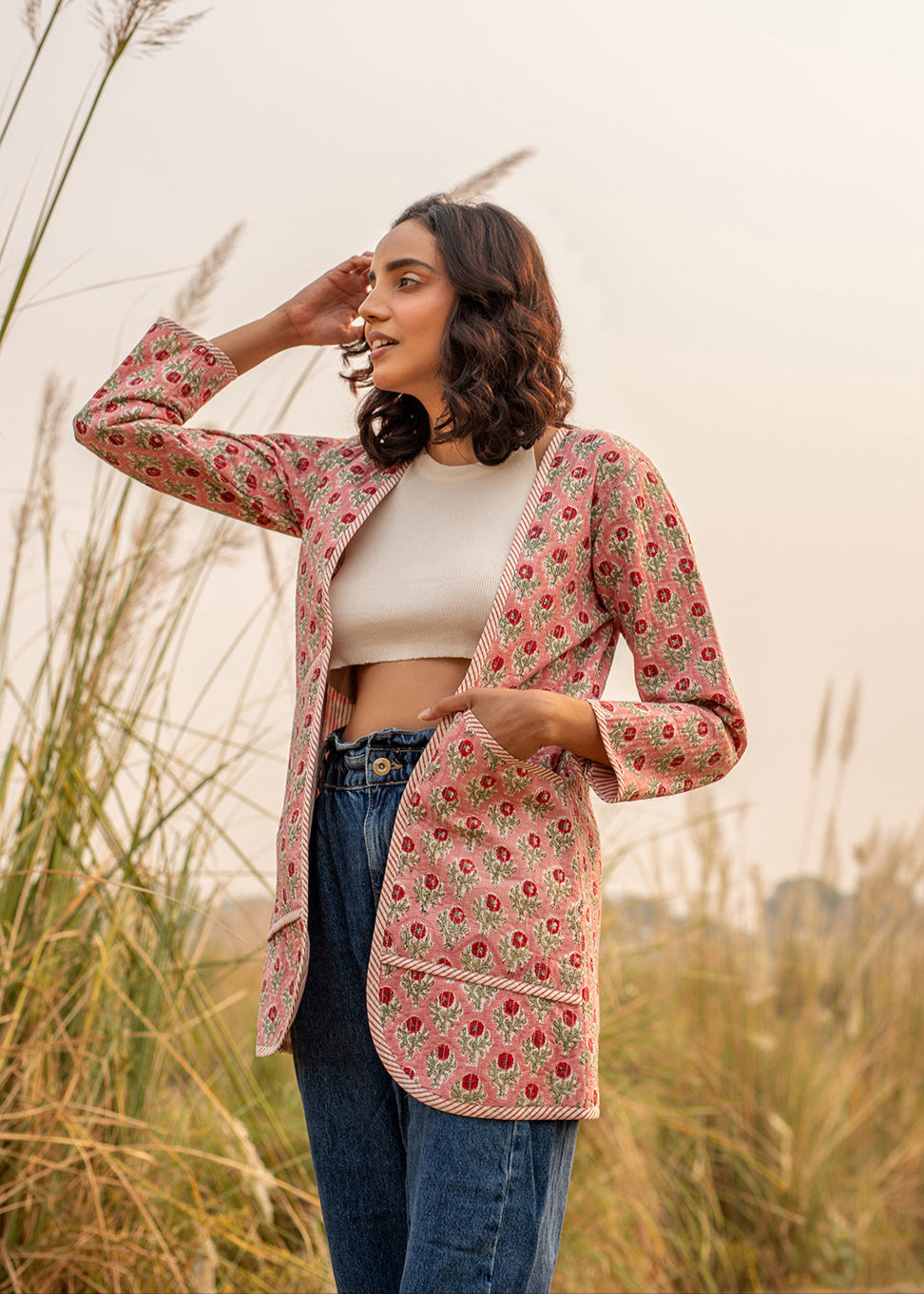 Woman wearing a floral hand block printed quilted jacket in pink, paired with blue high-waist jeans and a white crop top, standing in a sunlit dry grass field.