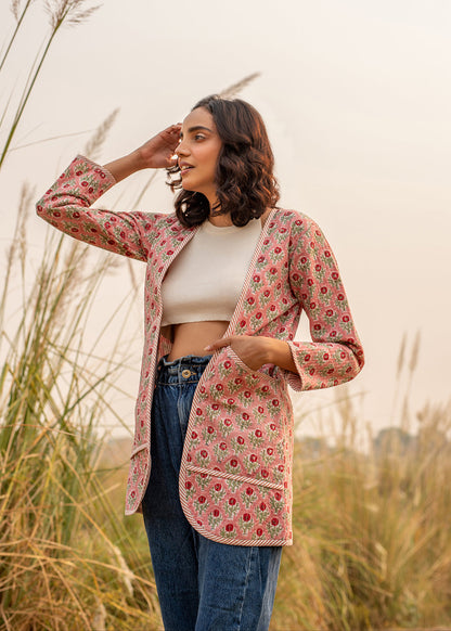 Woman wearing a floral hand block printed quilted jacket in pink, paired with blue high-waist jeans and a white crop top, standing in a sunlit dry grass field.