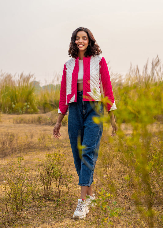 A young woman wearing a pink and white patchwork quilted jacket styled with high-waist blue jeans and a white crop top, smiling outdoors in a dry grass field.