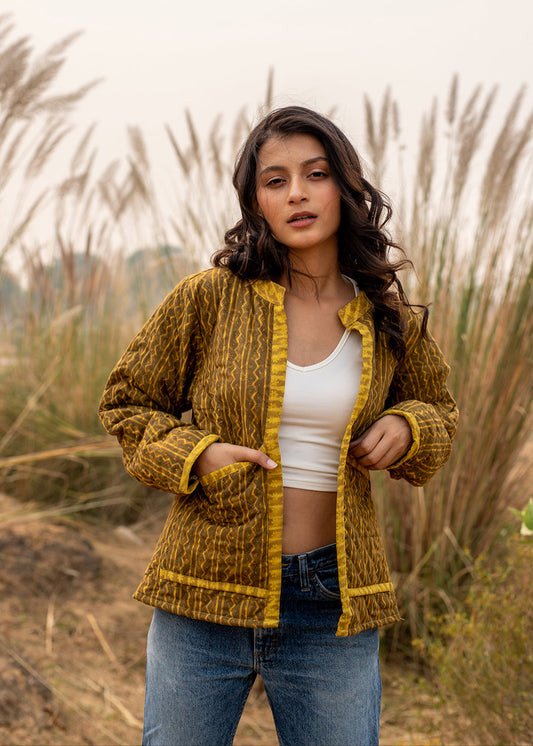 Woman wearing a mustard yellow patchwork jacket in a field, styled with a white crop top and jeans, arms open mid-twirl.
