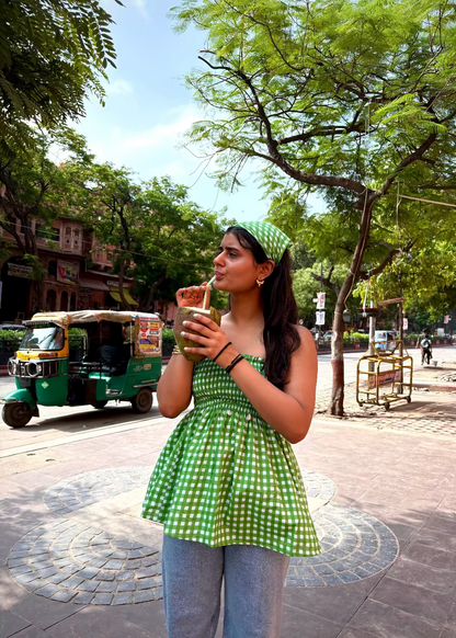 Woman in a green checkered LilacLoops - smocked tops That Romeo drinking from a cup on a street with trees and a rickshaw in the background.