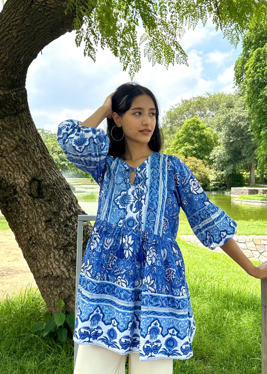Woman wearing a blue and white patterned Floral Whispers Tunic top That Romeo dress standing outdoors near a tree and body of water.
