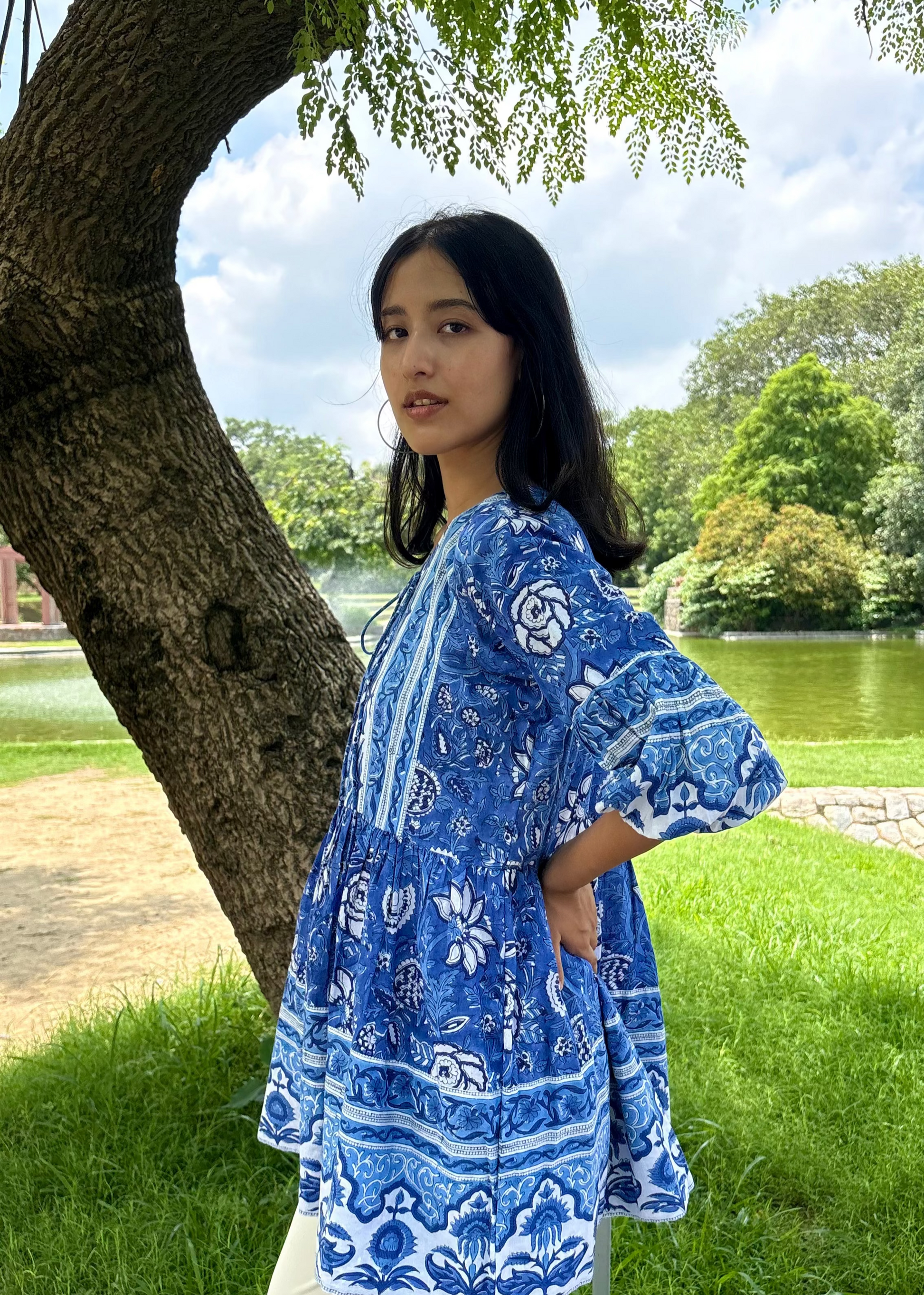 Woman wearing a blue and white patterned Floral Whispers Tunic top That Romeo dress standing outdoors near a tree and body of water.