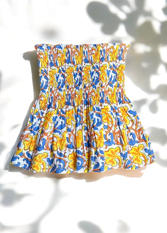 A strapless cotton smocked top with a bold yellow, blue, and orange baroque floral print, flared below the bust and laid flat under soft shadows by That Romeo