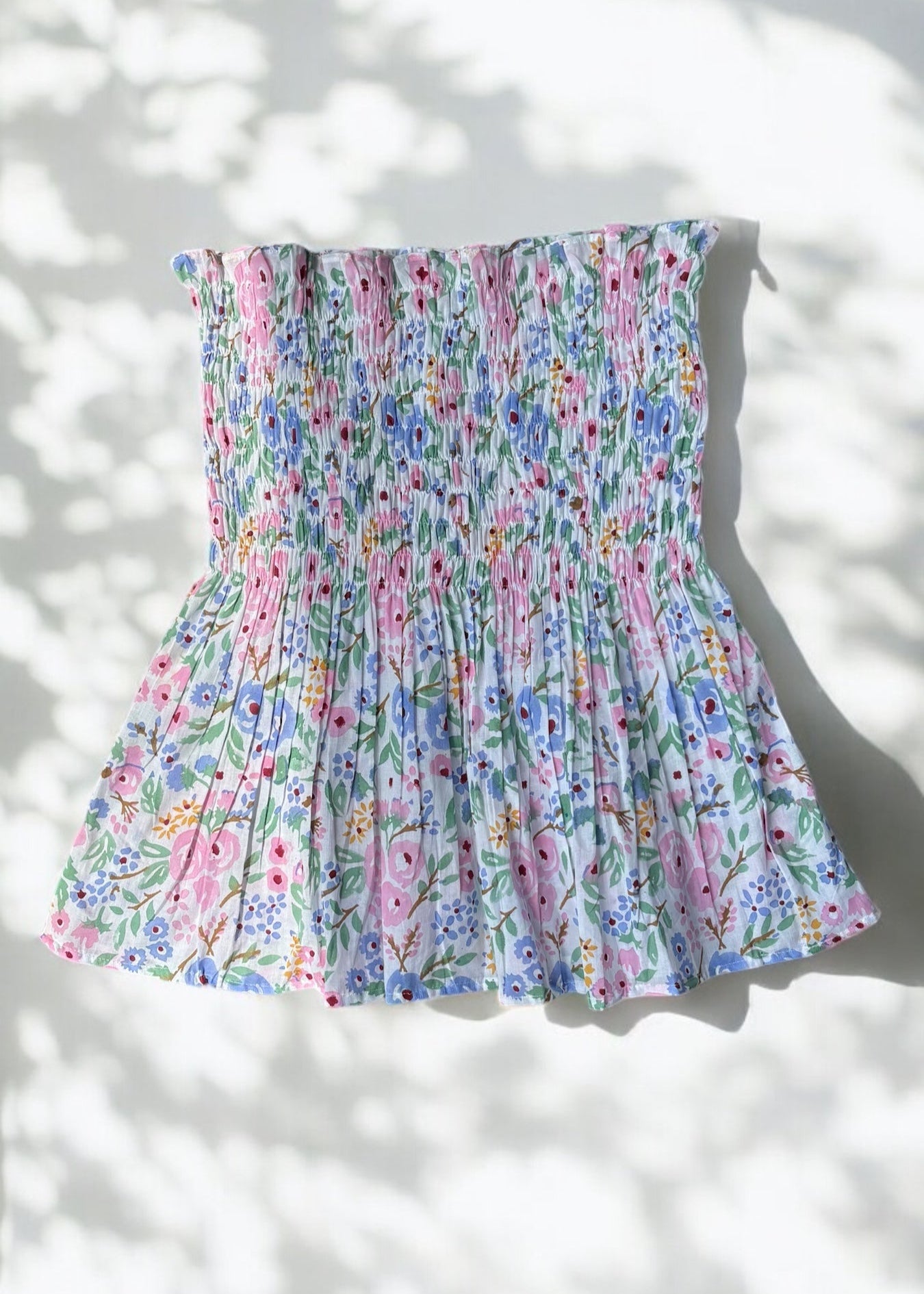 A strapless floral smocked top in pastel shades of pink, blue, and green, flared below the bust and photographed against a softly lit wall with leafy shadows by That Romeo