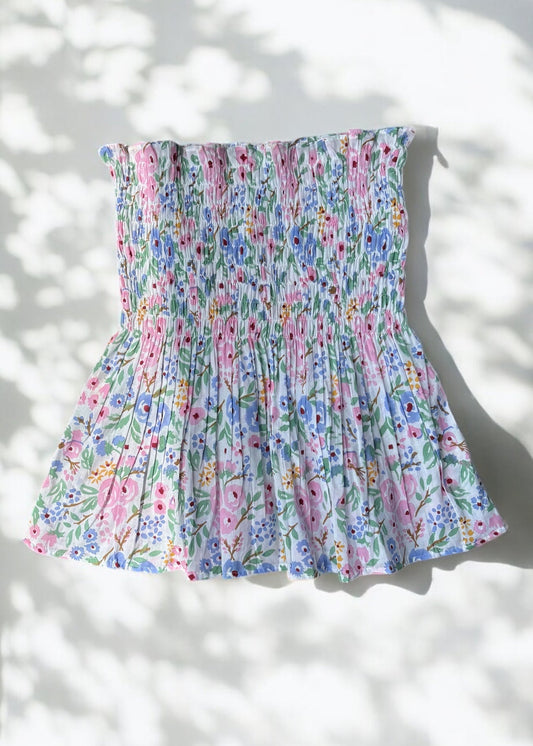 A strapless floral smocked top in pastel shades of pink, blue, and green, flared below the bust and photographed against a softly lit wall with leafy shadows by That Romeo