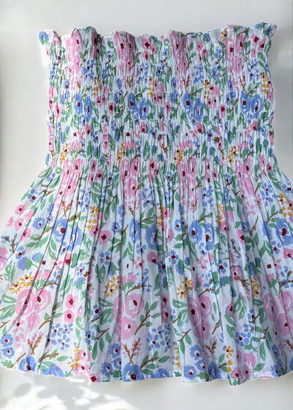 A strapless floral smocked top in pastel shades of pink, blue, and green, flared below the bust and photographed against a softly lit wall with leafy shadows by That Romeo