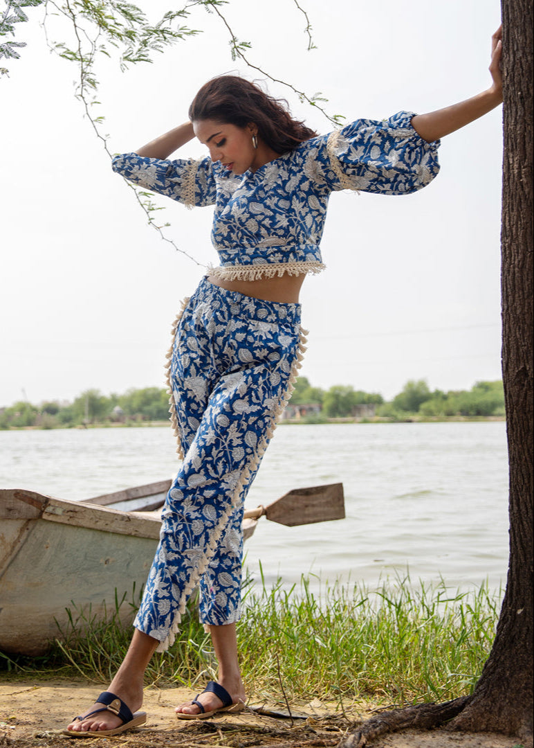 Woman wearing a blue floral hand block printed cotton co-ord set with tassel pants and a fringe-trimmed crop top, standing beside a tree near a lake.