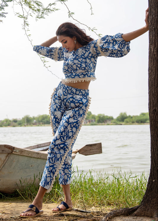 Woman wearing a blue floral hand block printed cotton co-ord set with tassel pants and a fringe-trimmed crop top, standing beside a tree near a lake.