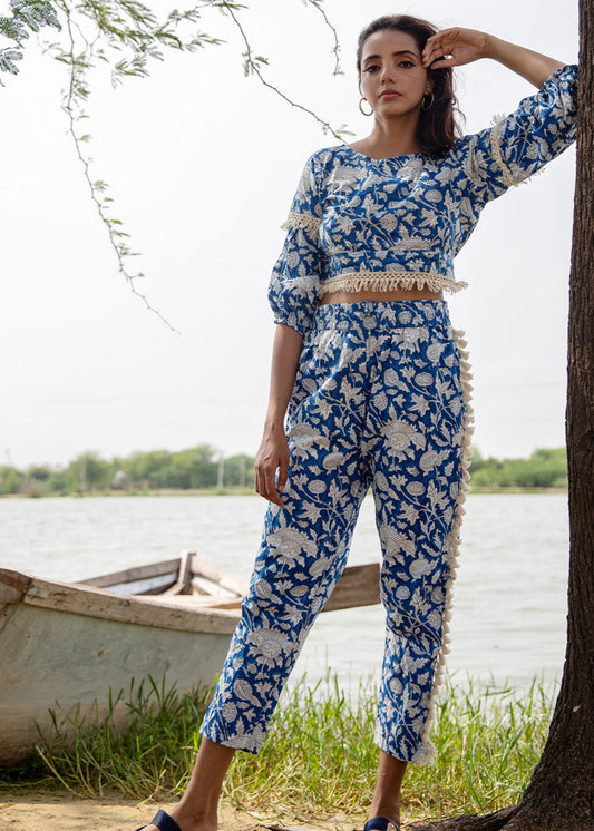 Woman wearing a blue floral hand block printed cotton co-ord set with tassel pants and a fringe-trimmed crop top, standing beside a tree near a lake.