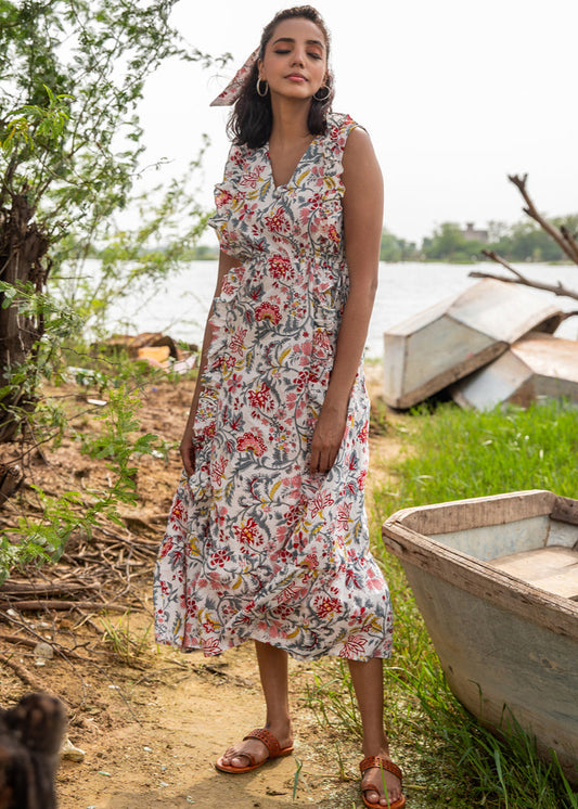 Woman wearing a sleeveless floral handblock printed cotton maxi dress with frill neckline and waist tie, standing outdoors near a rustic boat and greenery.