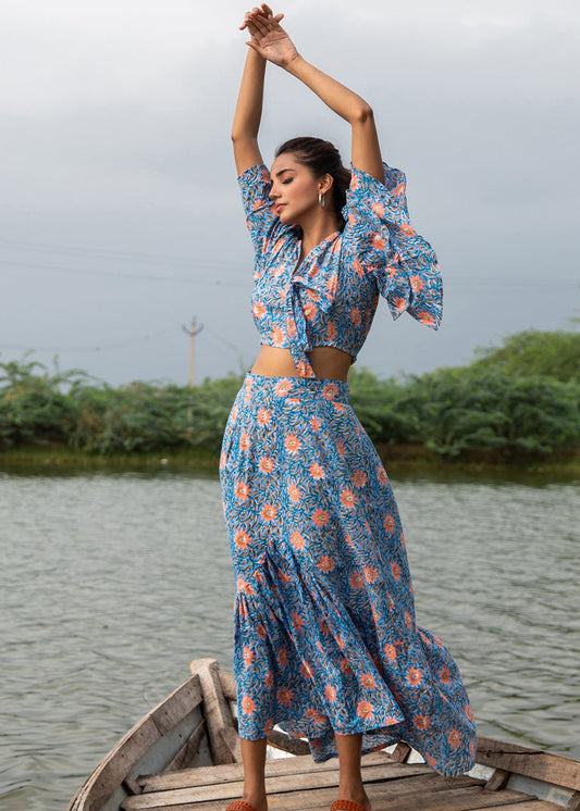 Woman wearing a floral hand block printed crop top and flared skirt co-ord set with frilled sleeves, standing on a wooden boat with arms raised, beside a serene lake.

