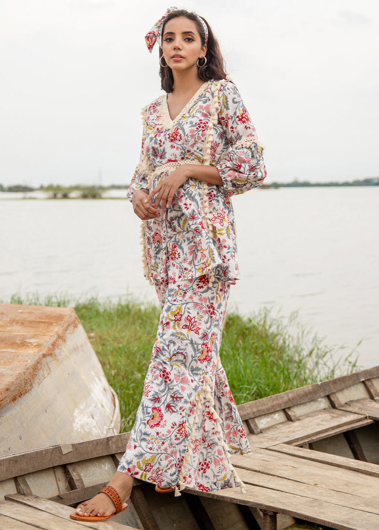 A woman wearing a hand block printed cotton co-ord set with a bohemian short kurta and flared pants, styled with fringe and tassel details, standing by the water on a wooden boat.