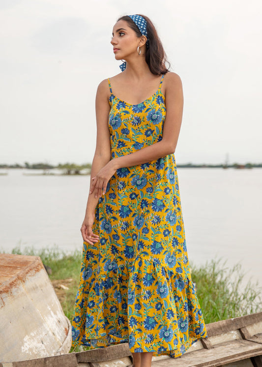 Woman wearing a yellow floral block printed cotton maxi dress with blue patterns and spaghetti straps, standing near a lake on a wooden boat, exuding breezy summer vibes.