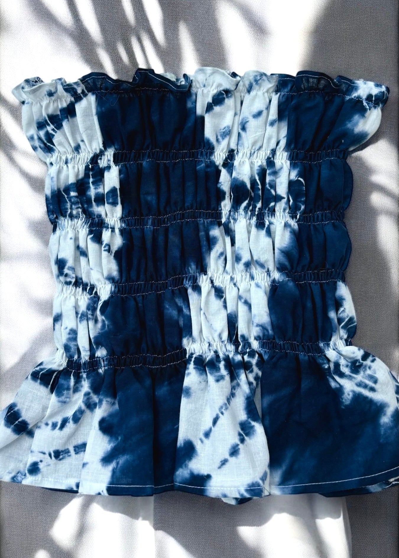 Flat lay of Shibori-dyed indigo and white smocked cotton bust top with ruffled hem, captured in natural sunlight and shadow play.