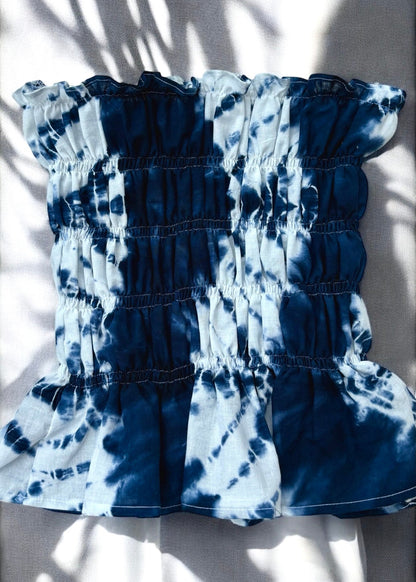 Flat lay of Shibori-dyed indigo and white smocked cotton bust top with ruffled hem, captured in natural sunlight and shadow play.