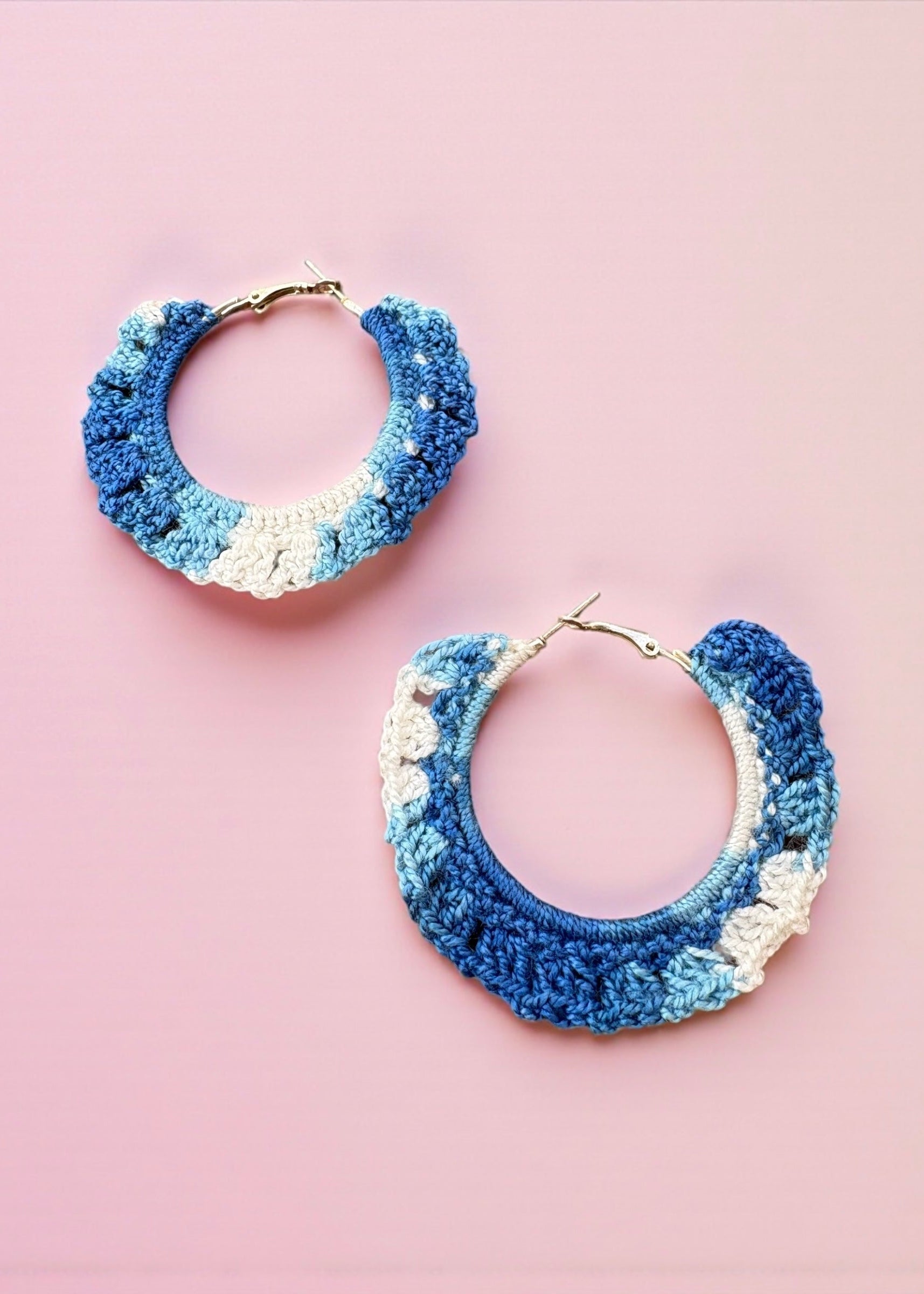 Hand-crocheted blue and ivory hoop earrings by ThatRomeo on a pink background.