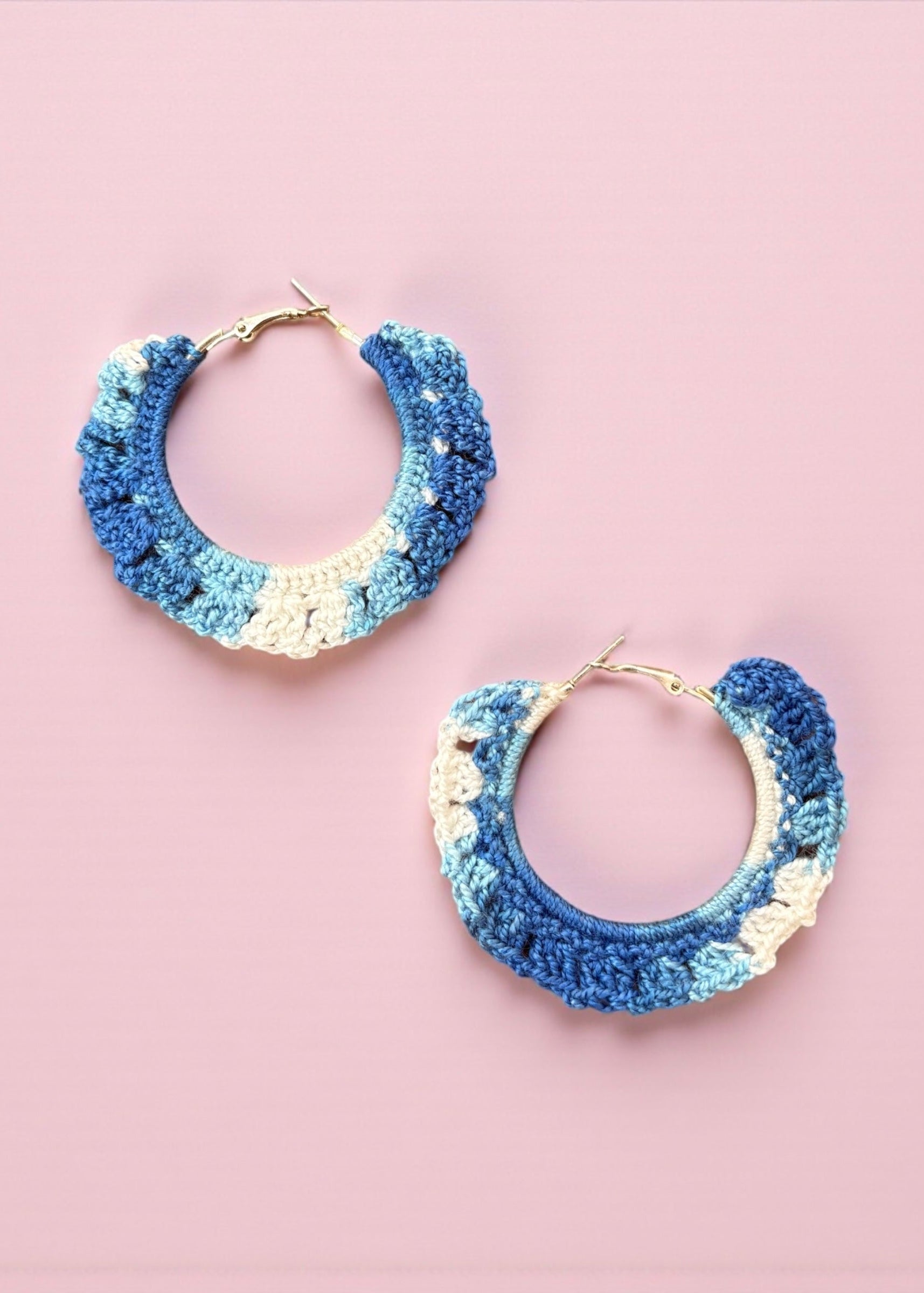 Hand-crocheted blue and ivory hoop earrings by ThatRomeo on a pink background.