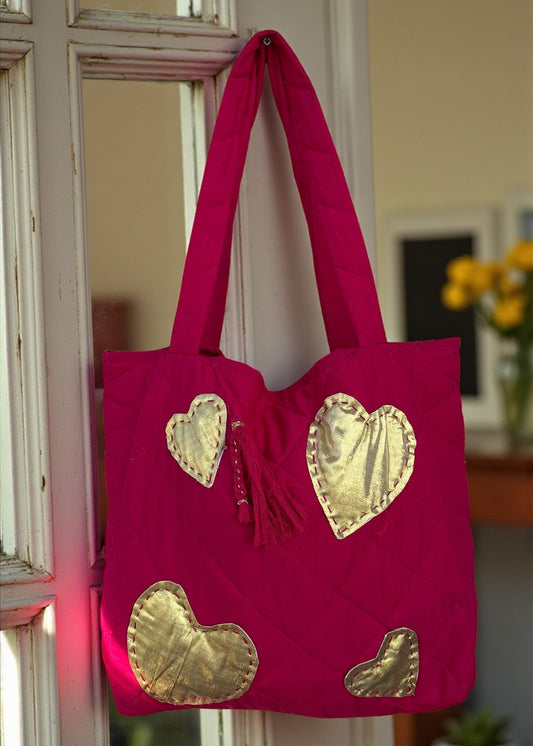 Fuchsia quilted tote bag with large gold heart appliqués and a matching tassel charm hanging by a window.