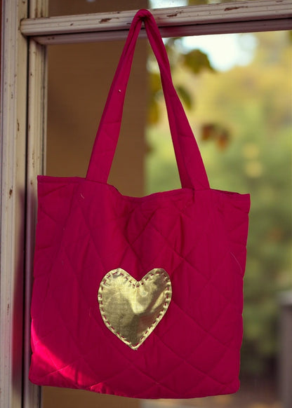 Fuchsia quilted tote bag with large gold heart appliqués and a matching tassel charm hanging by a window.