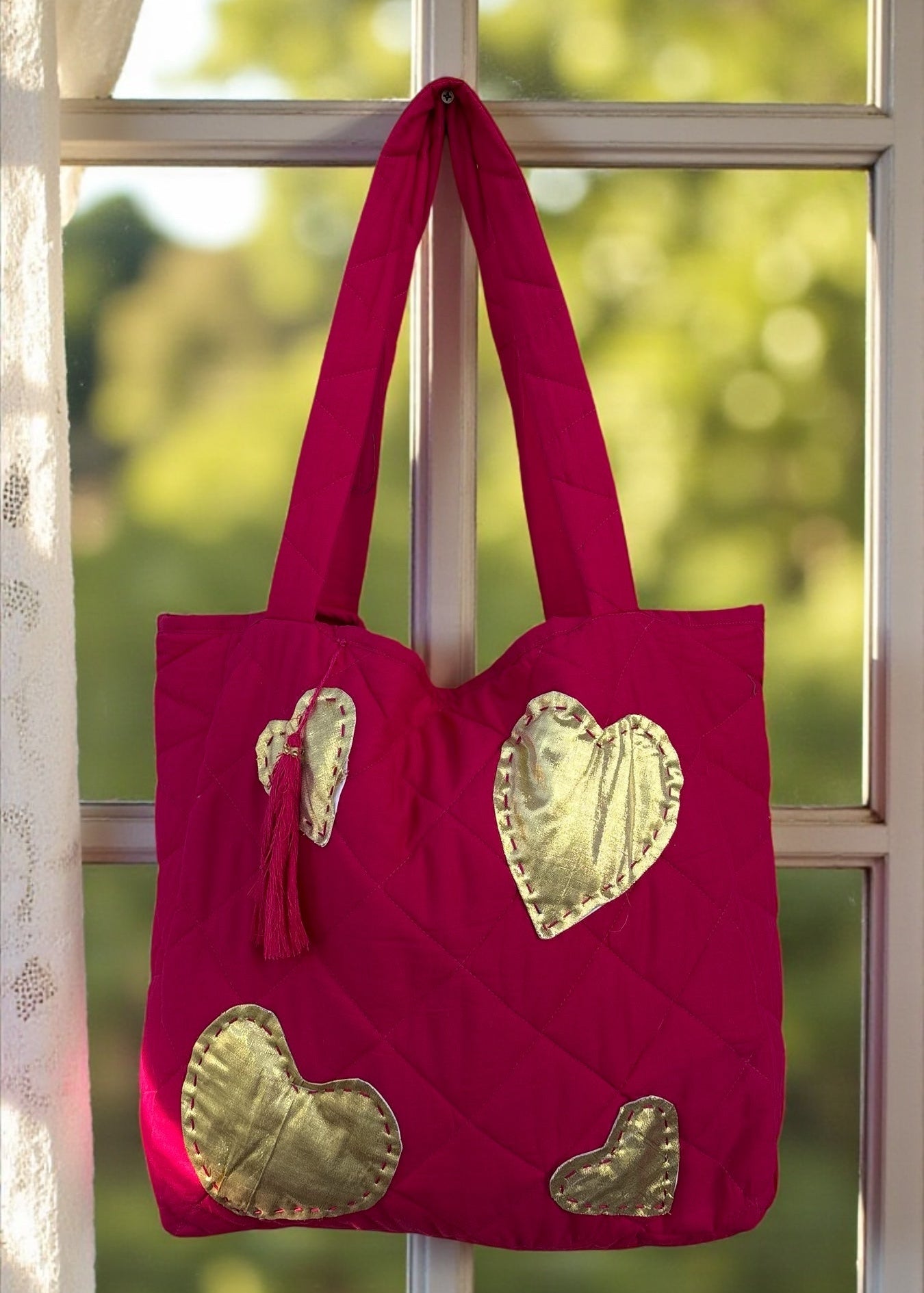 Fuchsia quilted tote bag with large gold heart appliqués and a matching tassel charm hanging by a window.