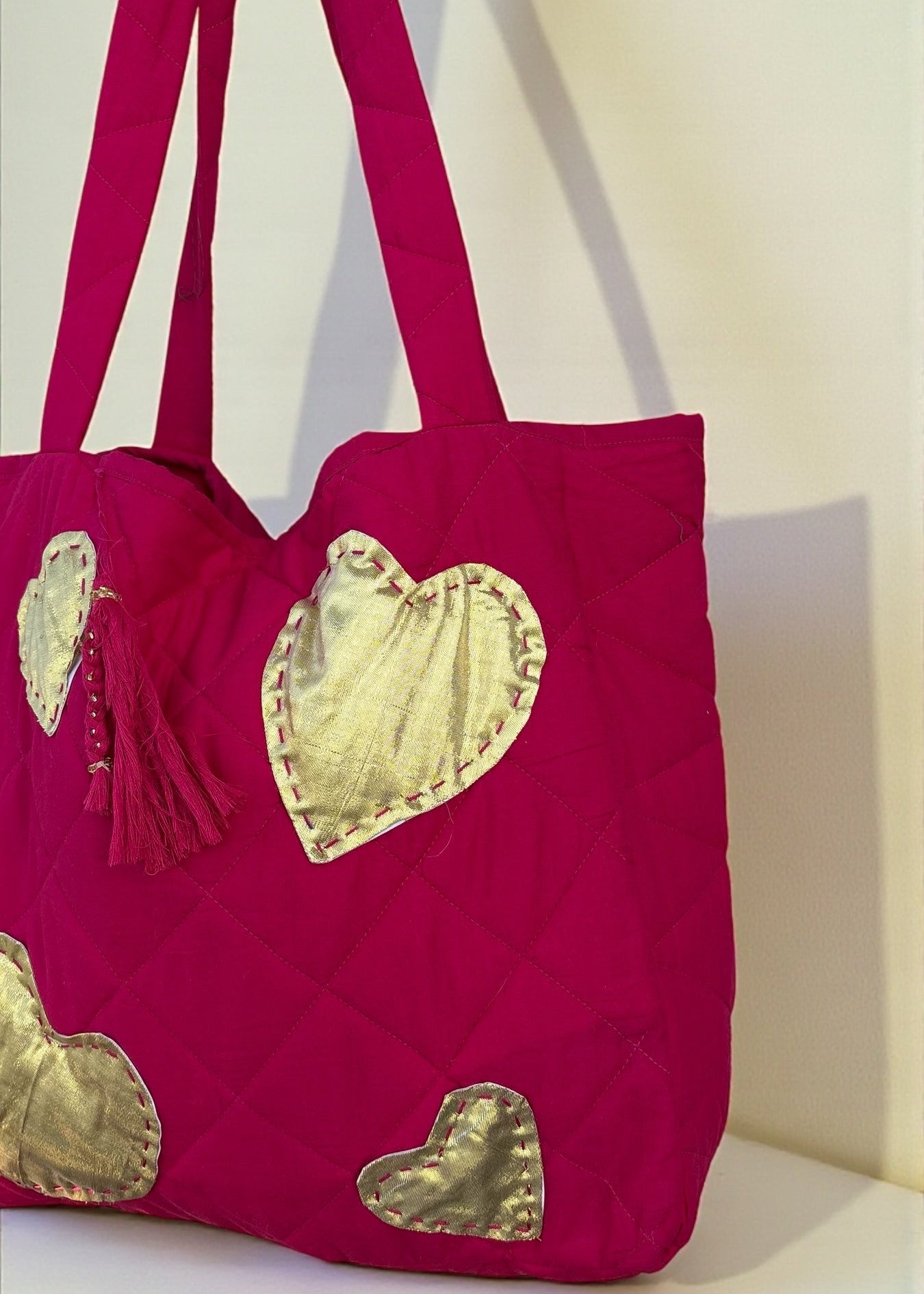 Fuchsia quilted tote bag with large gold heart appliqués and a matching tassel charm hanging by a window.