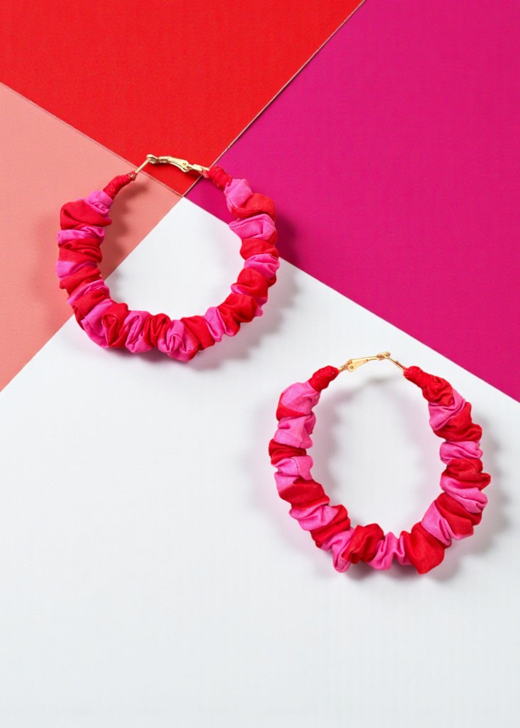 Pink and red ruched fabric hoop earrings by ThatRomeo on a colour-blocked background.