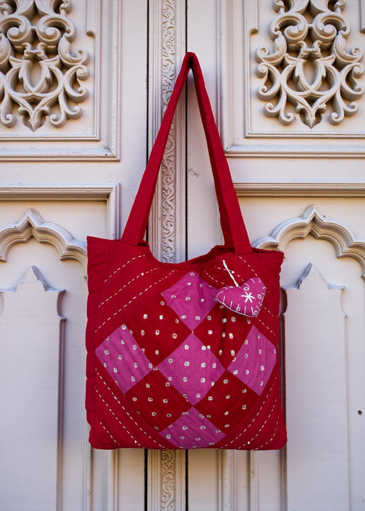 Red quilted cotton tote bag with pink patchwork panel and heart charm hanging on a carved white door.