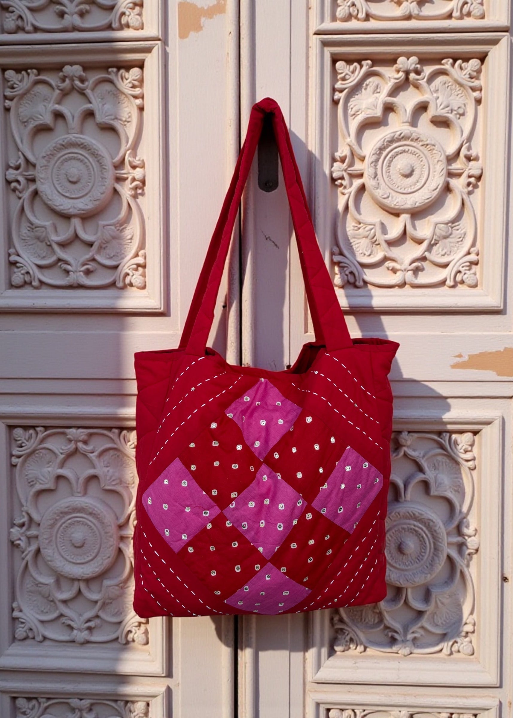 Red quilted cotton tote bag with pink patchwork panel and heart charm hanging on a carved white door.