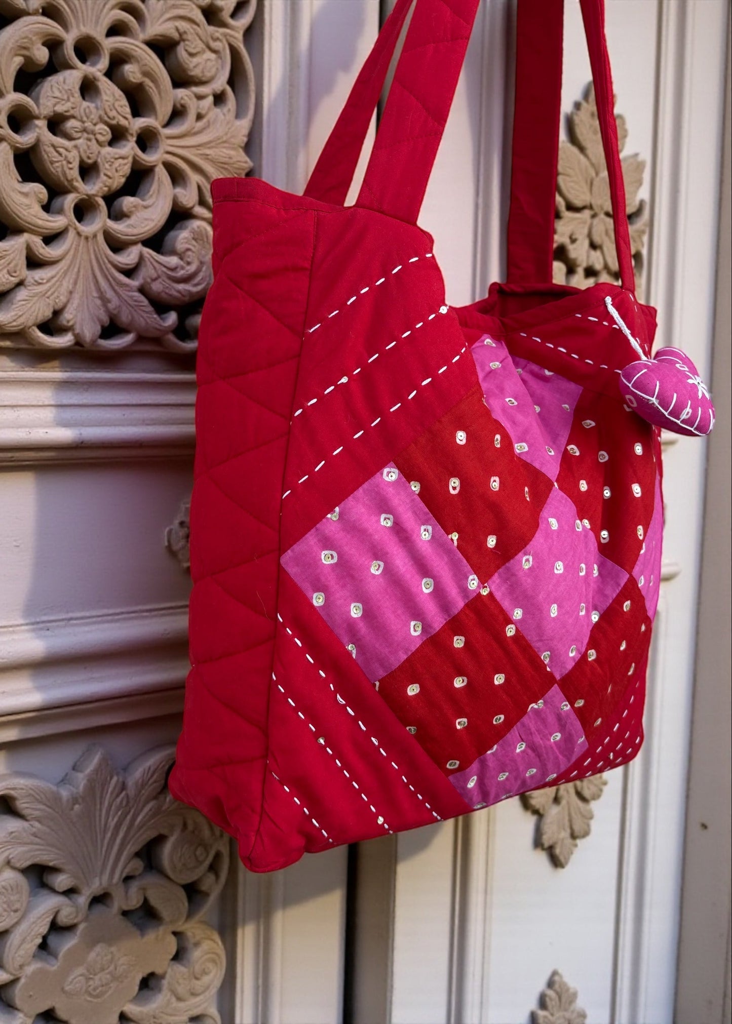 Red quilted cotton tote bag with pink patchwork panel and heart charm hanging on a carved white door.