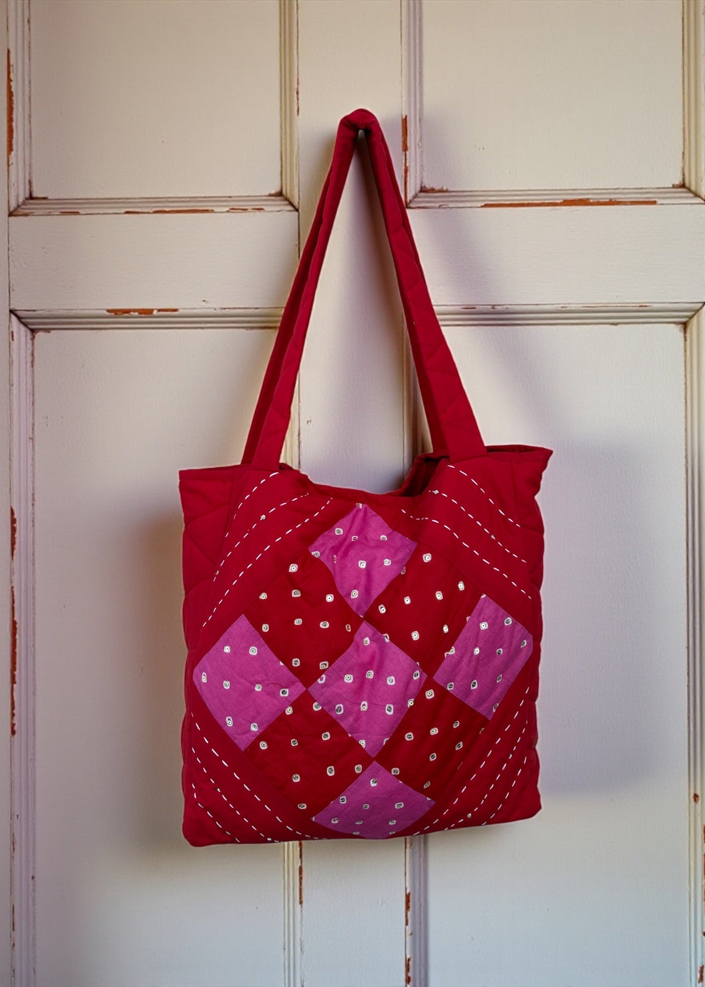 Red quilted cotton tote bag with pink patchwork panel and heart charm hanging on a carved white door.