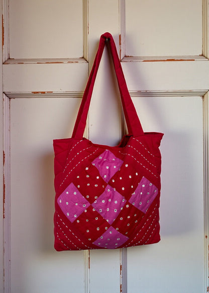 Red quilted cotton tote bag with pink patchwork panel and heart charm hanging on a carved white door.