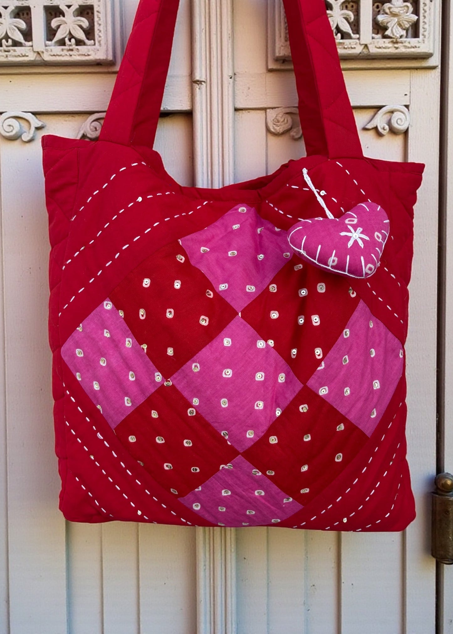 Red quilted cotton tote bag with pink patchwork panel and heart charm hanging on a carved white door.