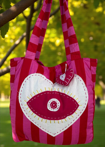 Pink and red striped quilted tote bag with an oversized heart appliqué and evil-eye embroidery, hanging outdoors.