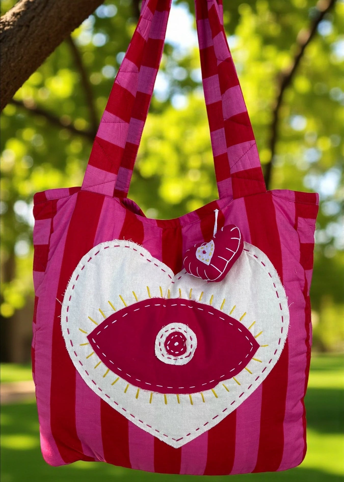 Pink and red striped quilted tote bag with an oversized heart appliqué and evil-eye embroidery, hanging outdoors.
