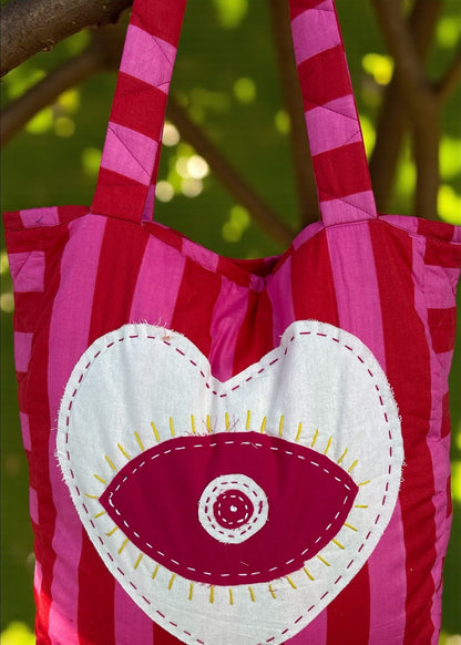 Pink and red striped quilted tote bag with an oversized heart appliqué and evil-eye embroidery, hanging outdoors.