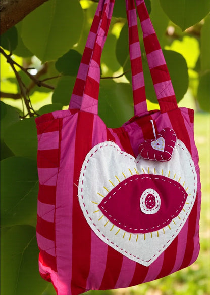 Pink and red striped quilted tote bag with an oversized heart appliqué and evil-eye embroidery, hanging outdoors.