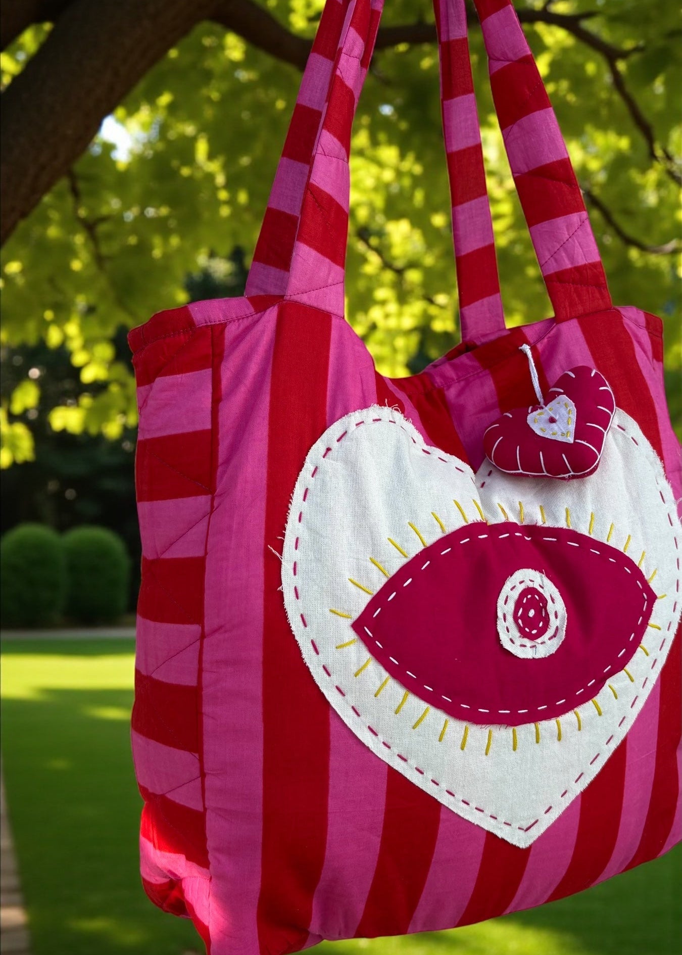 Pink and red striped quilted tote bag with an oversized heart appliqué and evil-eye embroidery, hanging outdoors.
