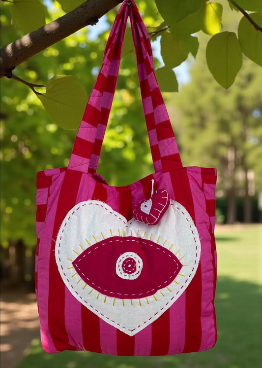 Pink and red striped quilted tote bag with an oversized heart appliqué and evil-eye embroidery, hanging outdoors.