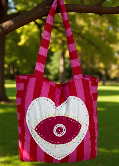 Pink and red striped quilted tote bag with an oversized heart appliqué and evil-eye embroidery, hanging outdoors.