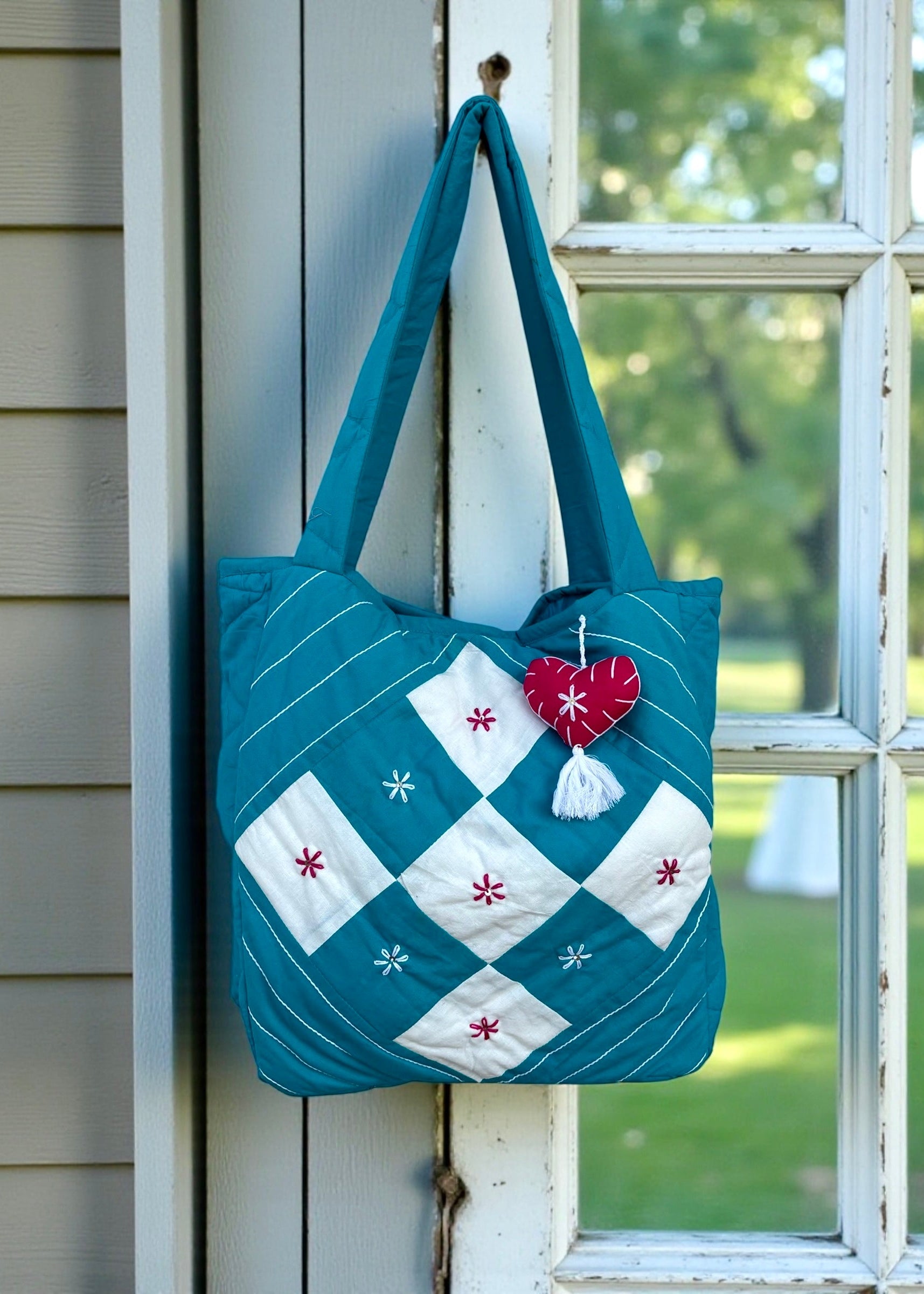 Teal quilted cotton tote bag with white patchwork panel and embroidered floral motifs hanging on a window frame.
