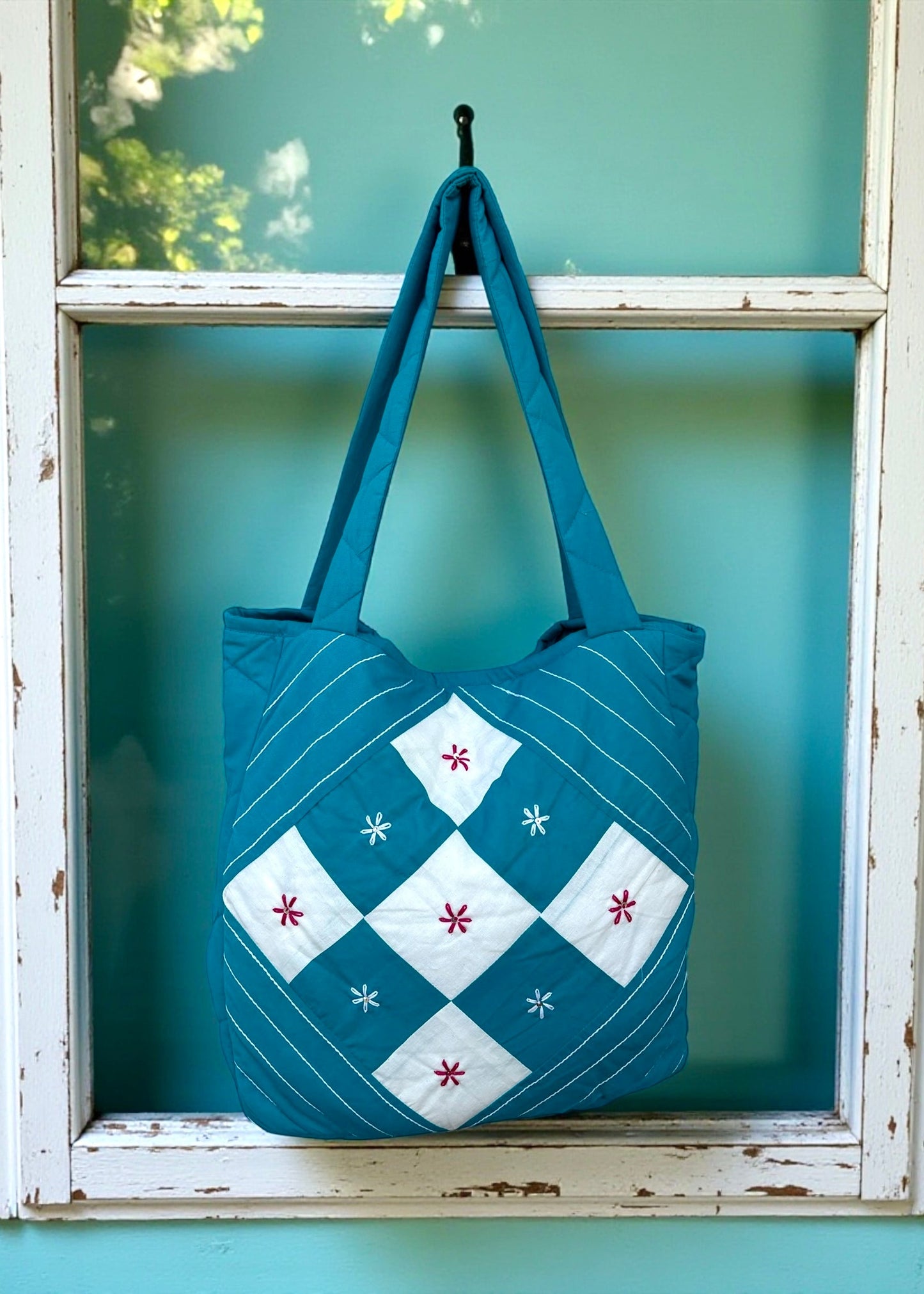 Teal quilted cotton tote bag with white patchwork panel and embroidered floral motifs hanging on a window frame.