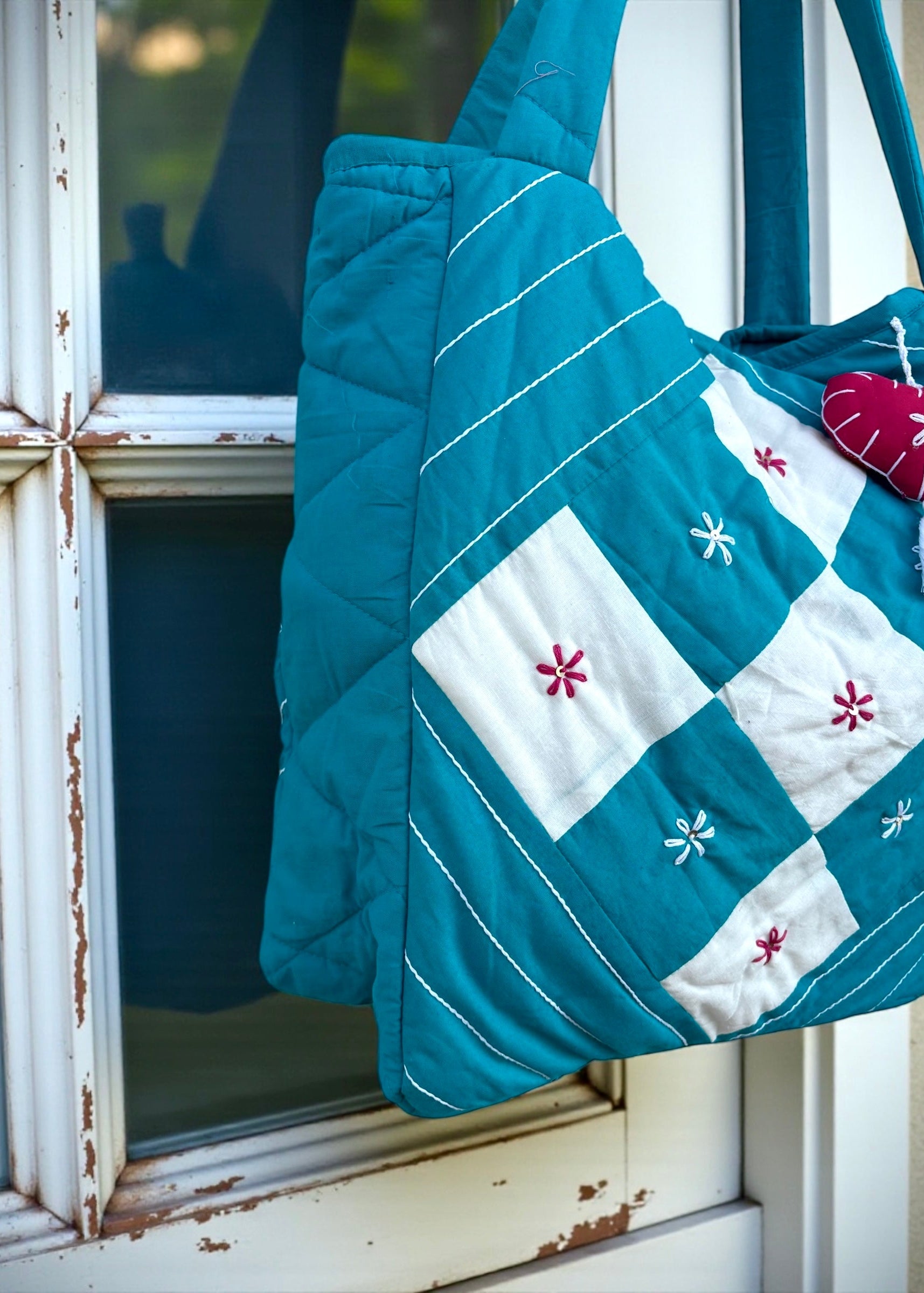 Teal quilted cotton tote bag with white patchwork panel and embroidered floral motifs hanging on a window frame.
