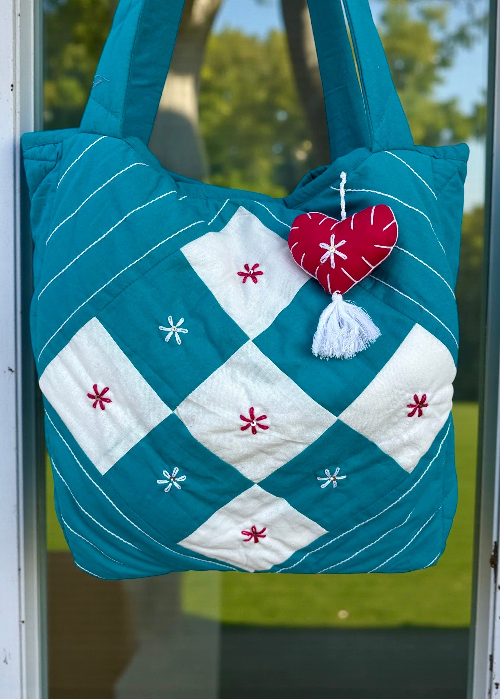 Teal quilted cotton tote bag with white patchwork panel and embroidered floral motifs hanging on a window frame.