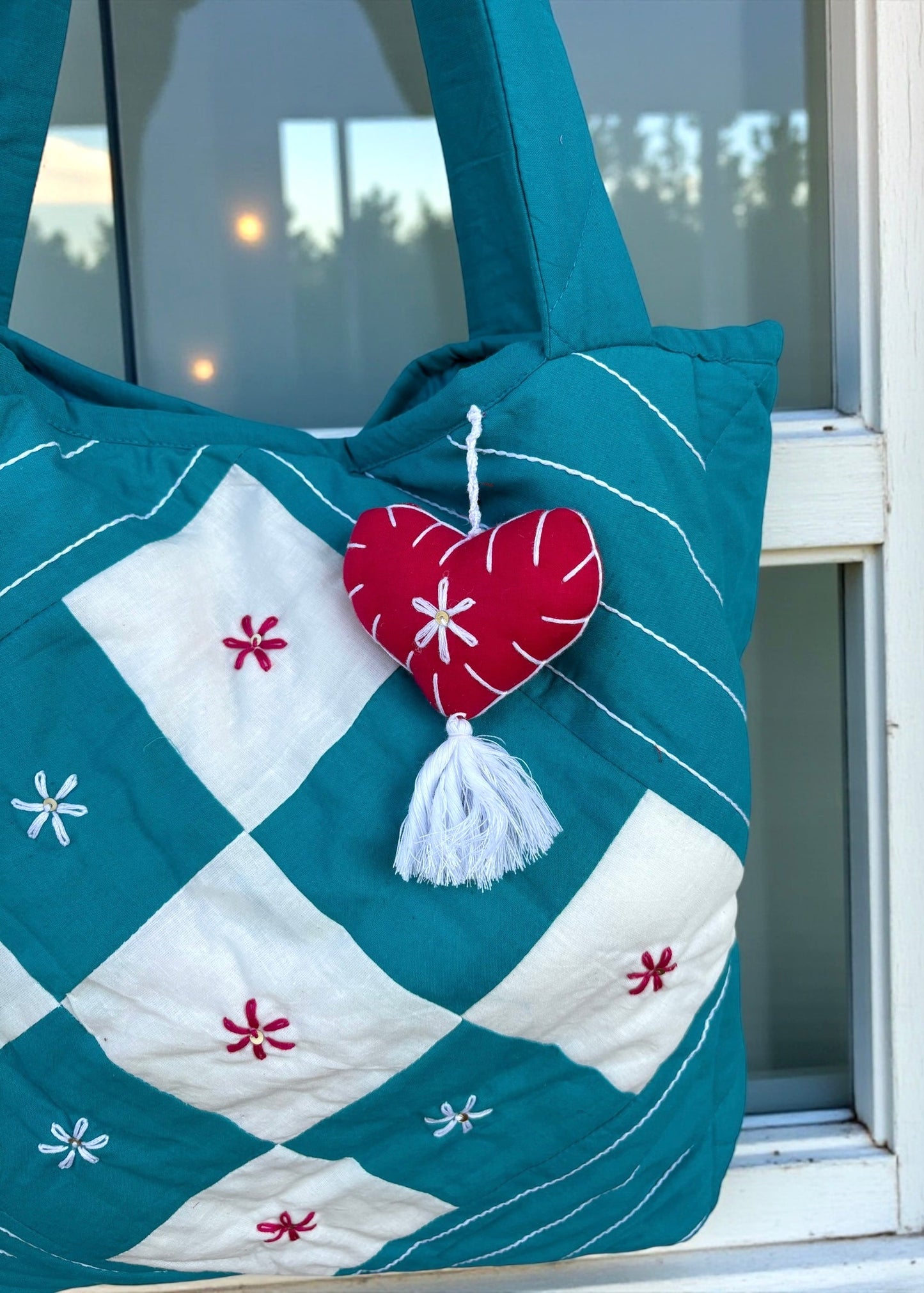 Teal quilted cotton tote bag with white patchwork panel and embroidered floral motifs hanging on a window frame.